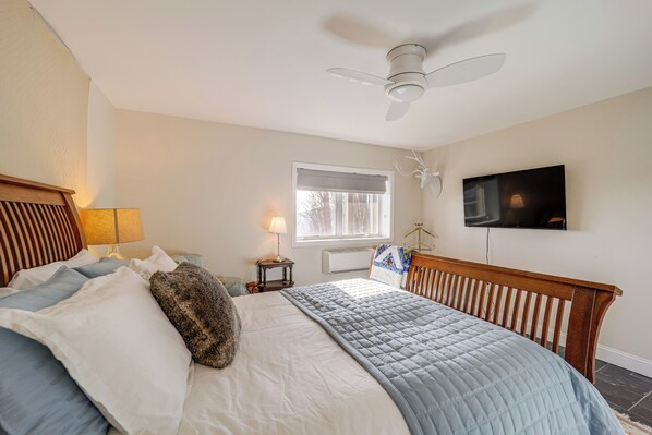 A bedroom with a bed, a ceiling fan, a window with blinds, a bedside table with a lamp, and a TV mounted on the wall.