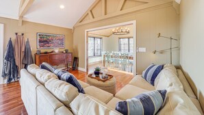 Ein geräumiges Wohnzimmer mit einer großen, beigefarbenen Eckcouch, Holzbalken an der Decke, einem Essbereich mit weißen Stühlen und einem großen Fenster, das natürliches Licht hereinlässt.