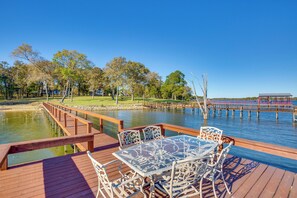 Een houten terras met een glazen tafel en stoelen, met uitzicht op een waterpartij, met een houten steiger die in het water uitloopt.