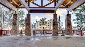 A covered outdoor area with stone pillars, a red railing, and a view of multi-story buildings in the background. - Walk to Ski Lift: Resort Condo in Park City! (Park City)