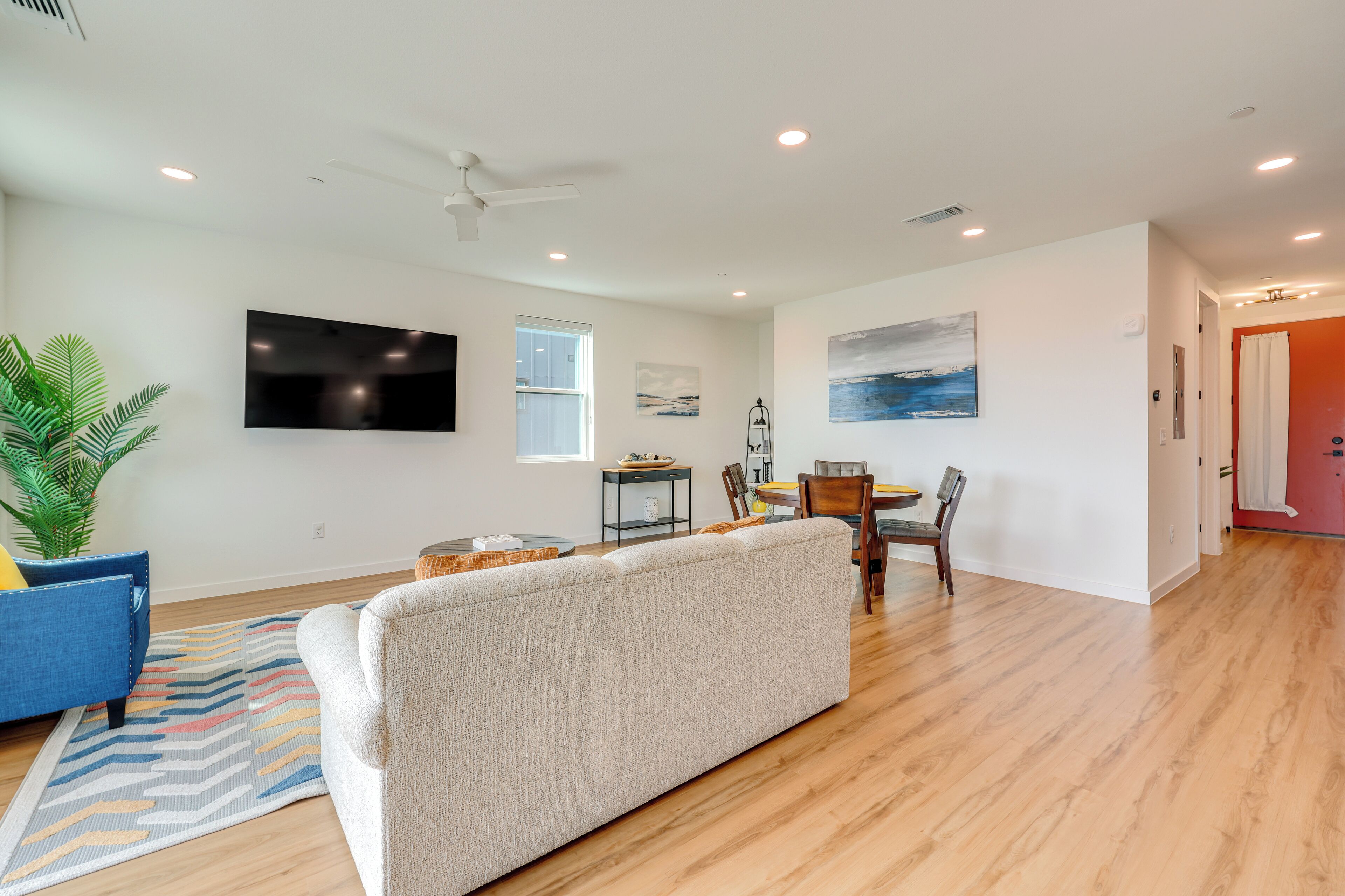 A modern living room with a large sofa, a dining area with a table and chairs, a flat-screen TV mounted on the wall, and a ceiling fan.