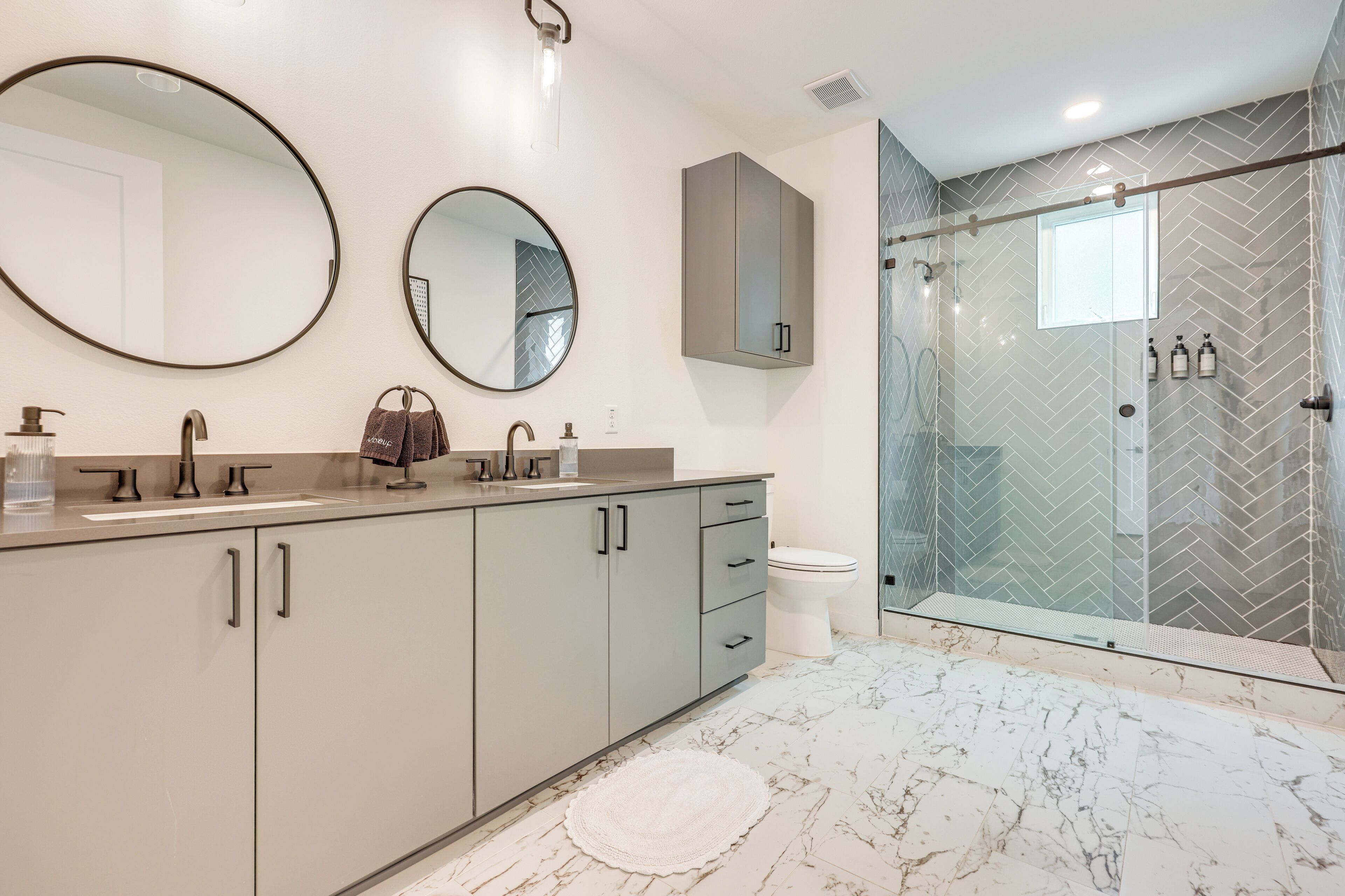 A modern bathroom with a marble floor, two round mirrors, a grey cabinet, a toilet, and a glass-enclosed shower.