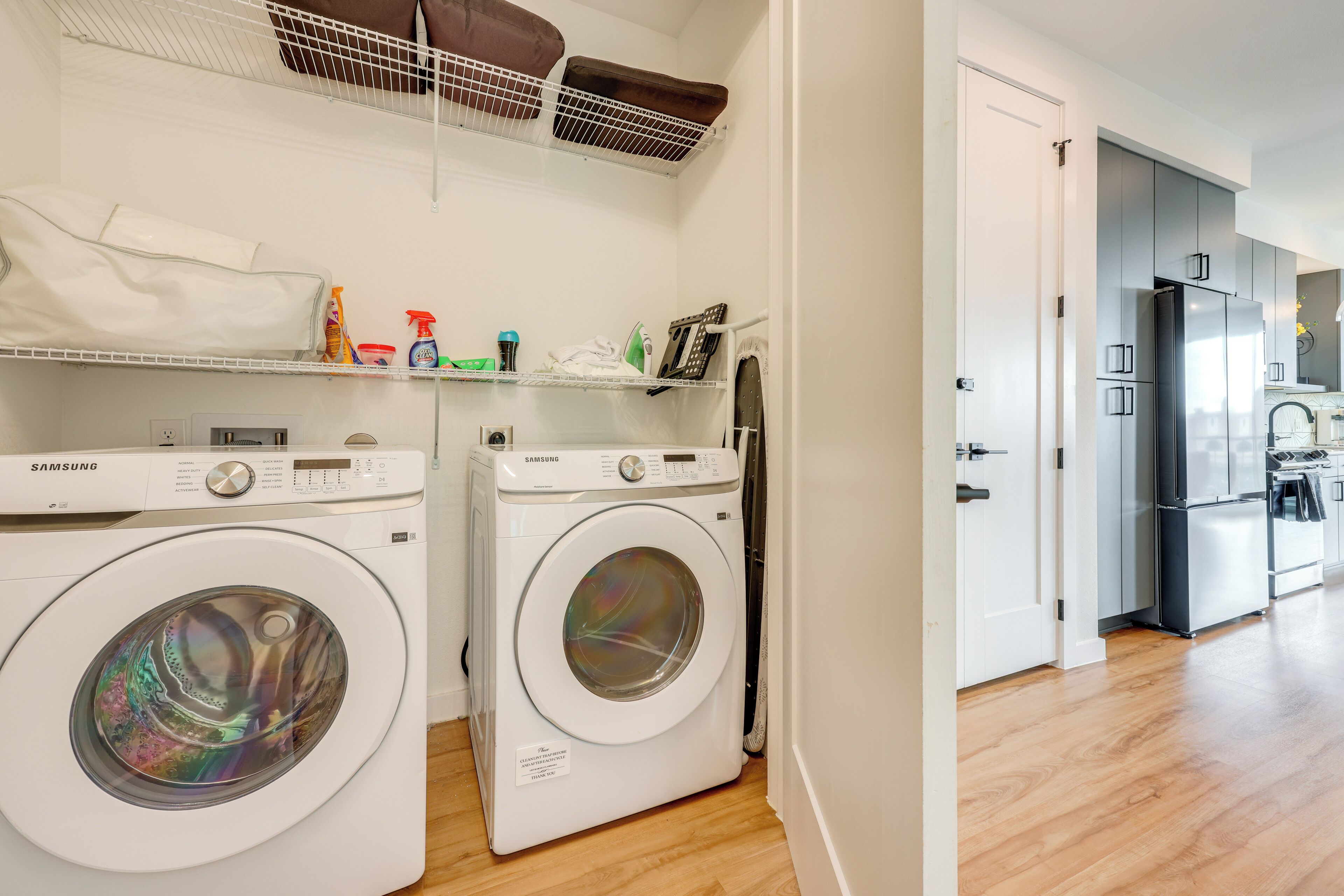 A laundry area with two Samsung washing machines, a drying rack, and a shelf with cleaning supplies.