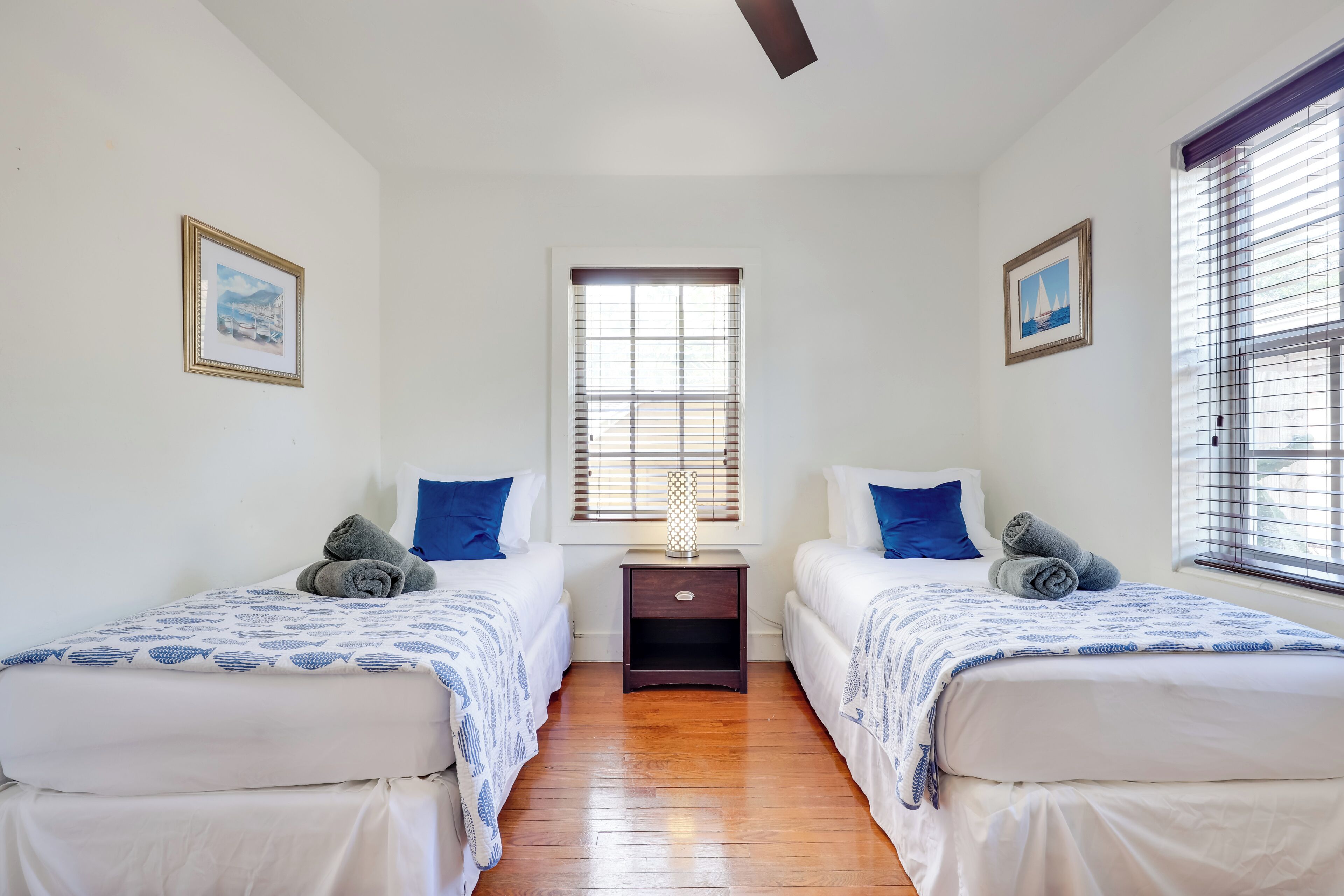 A hotel room with two beds, a nightstand, a window with blinds, and two framed pictures on the walls.