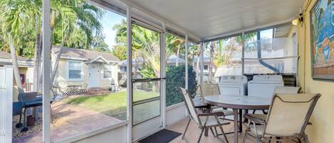 Un patio cubierto con una mesa y sillas, ventanas amplias y vistas a un jardín con palmeras y una casa.