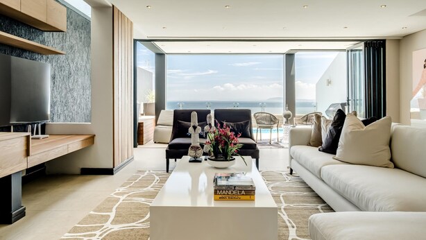 A modern living room with a large flat-screen TV, a white coffee table with books, a sofa, and a chair. The room has a large window offering a view of the sea. - Top Urban Escape w Pool and Ocean View Villa Alexa (Cape Town)