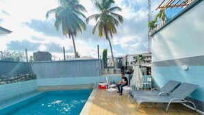 A pool area with a wooden deck, lounge chairs, and a person sitting by the pool. - HOUSE 10 SHORTLET  (Abuja)