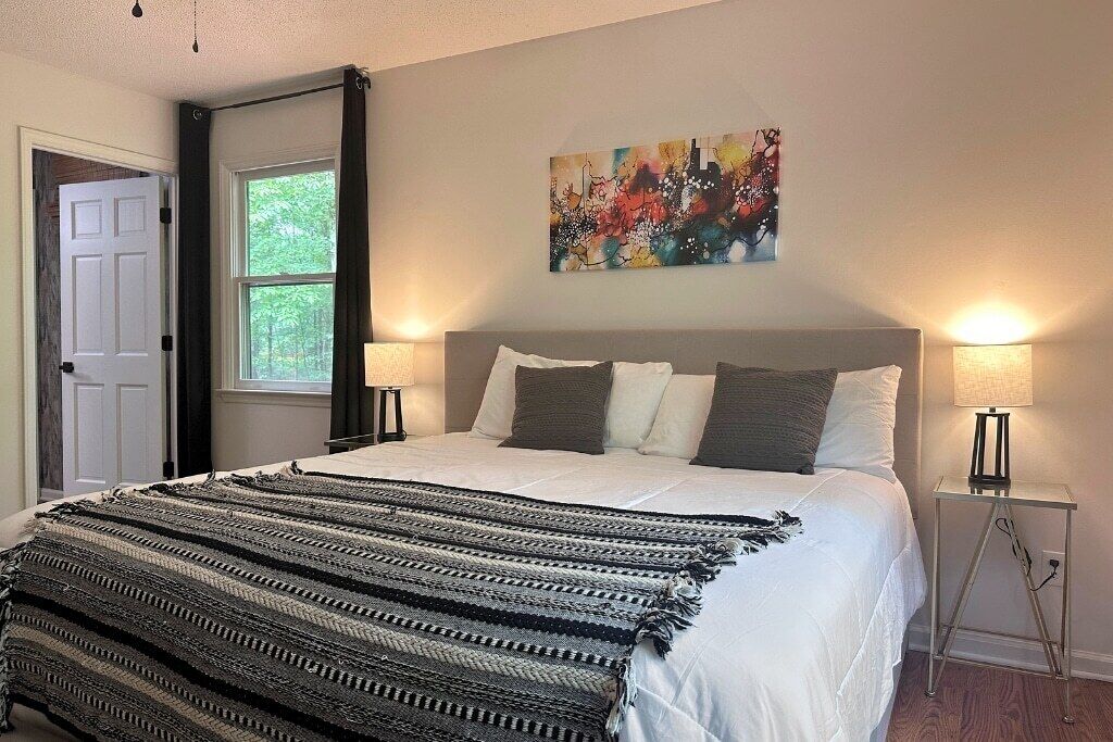 A bedroom with a bed, two bedside lamps, a window, a door, and a painting on the wall.