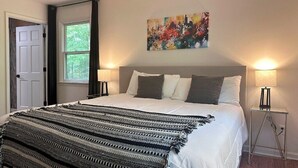 A bedroom with a bed, two bedside lamps, a window, a door, and a painting on the wall. - Modern Mountain at Rumbling Bald (Lake Lure)