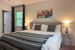 A bedroom with a bed, two bedside lamps, a window, a door, and a painting on the wall.