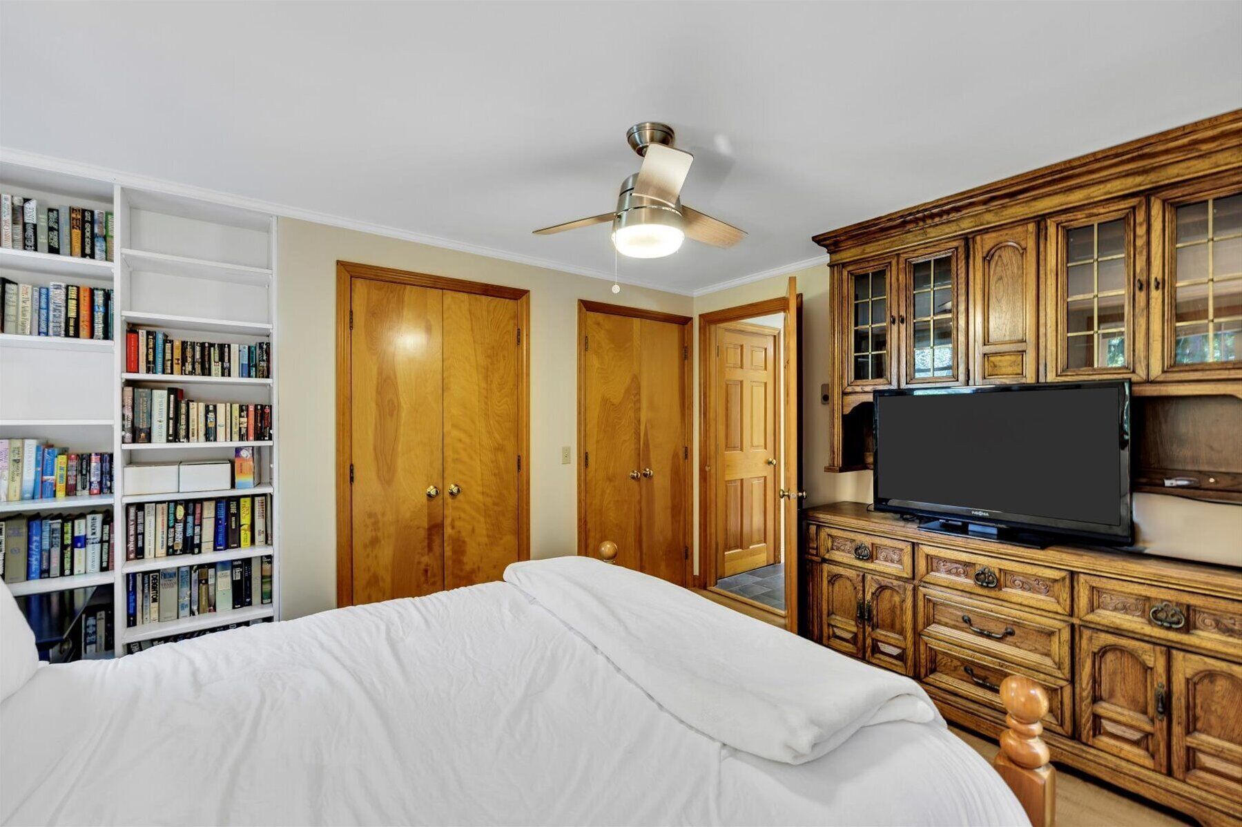 A bedroom with a bed, a bookshelf, a wardrobe, a ceiling fan, and a television.