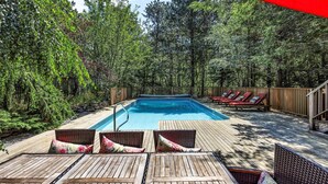 A swimming pool surrounded by wooden deck chairs and lounge chairs, with a wooden fence and dense foliage in the background. - Spacious Home With Private Heated Pool (2389) (Wellfleet)