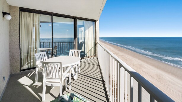 Varanda com mesa e cadeiras, com vista para uma praia e o oceano.