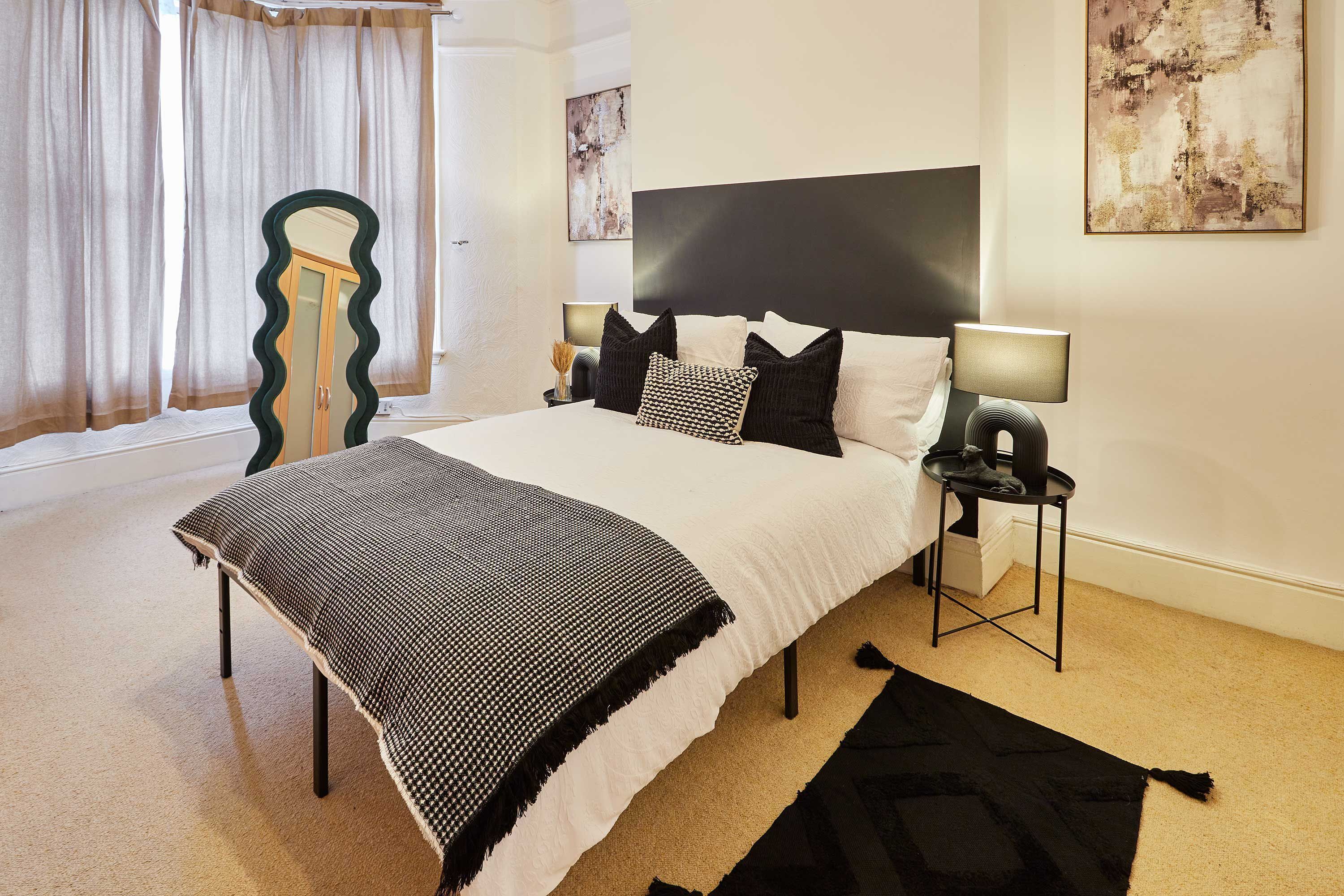 A bedroom with a large bed, a bedside table with a lamp, a mirror, and abstract wall art.