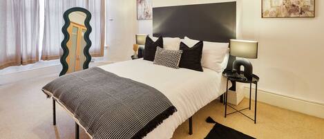A bedroom with a large bed, a bedside table with a lamp, a mirror, and abstract wall art.