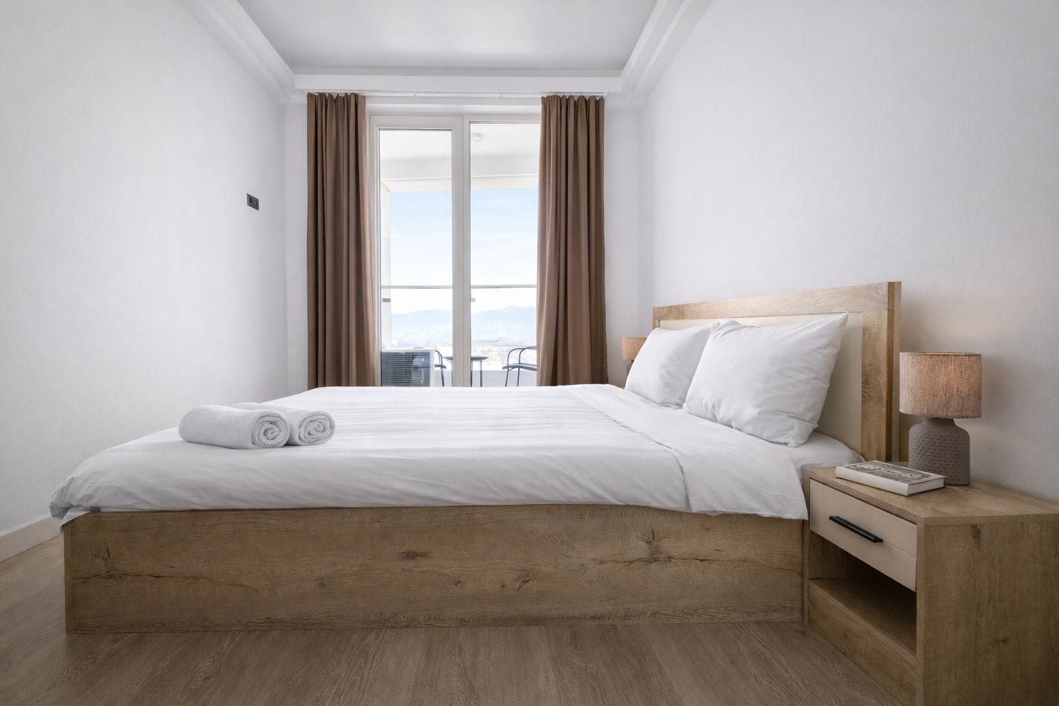 Habitación de hotel moderna con cama de madera, ropa de cama blanca, mesita de noche con lámpara y vistas al exterior a través de amplias ventanas.