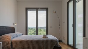 A bedroom with a bed, a nightstand, a floor lamp, and a large window with a view of greenery. - Loft 4 by Wonderful Italy (Olbia)