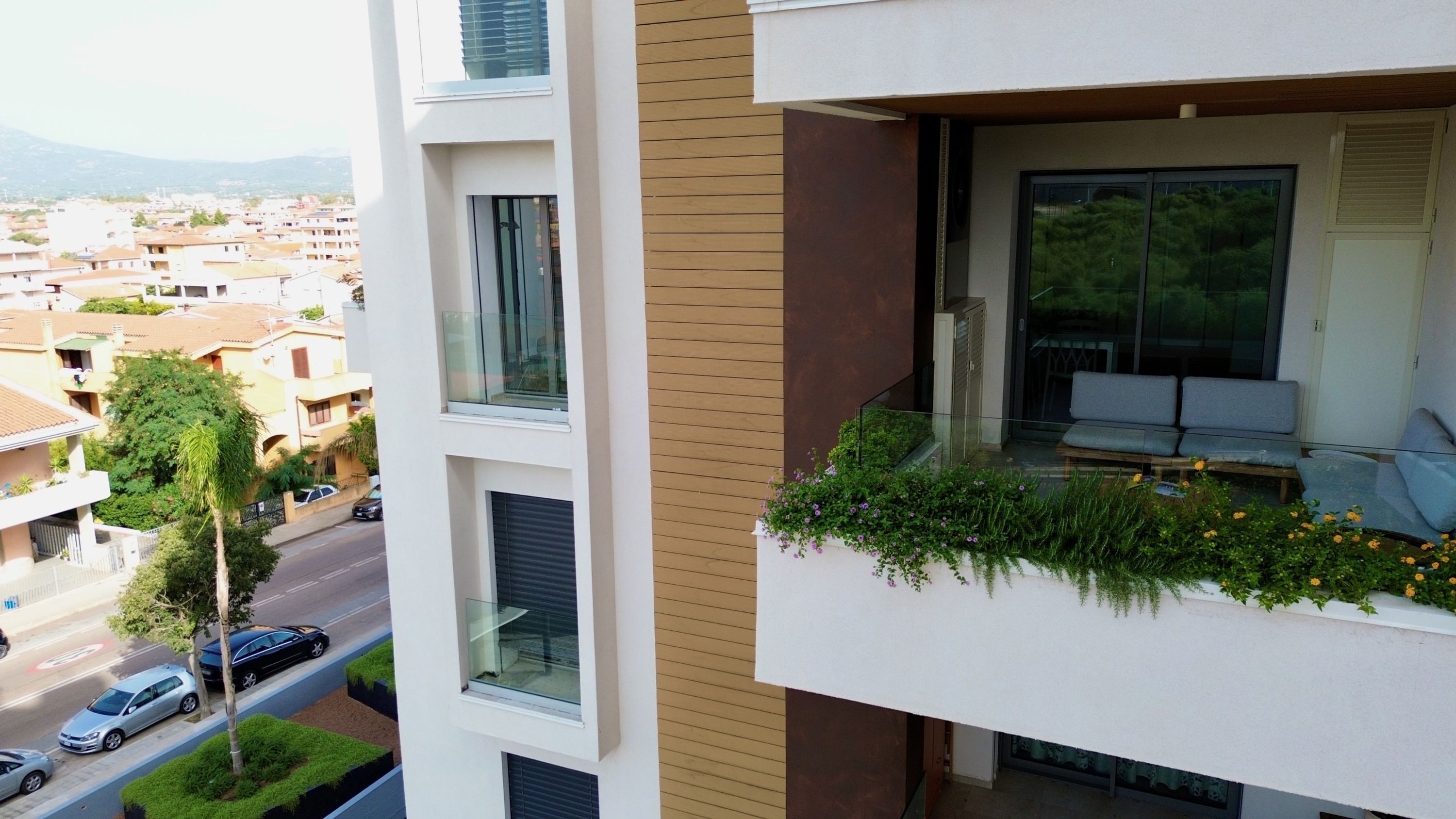 Um apartamento moderno com varanda, tendo guarda-corpo de vidro, área de estar com almofadas e vista para o bairro ao redor.