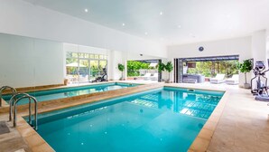 A modern indoor swimming pool with clear glass walls, a sun terrace with outdoor seating, and exercise equipment. - Host Stay The Grove (Hartlepool)