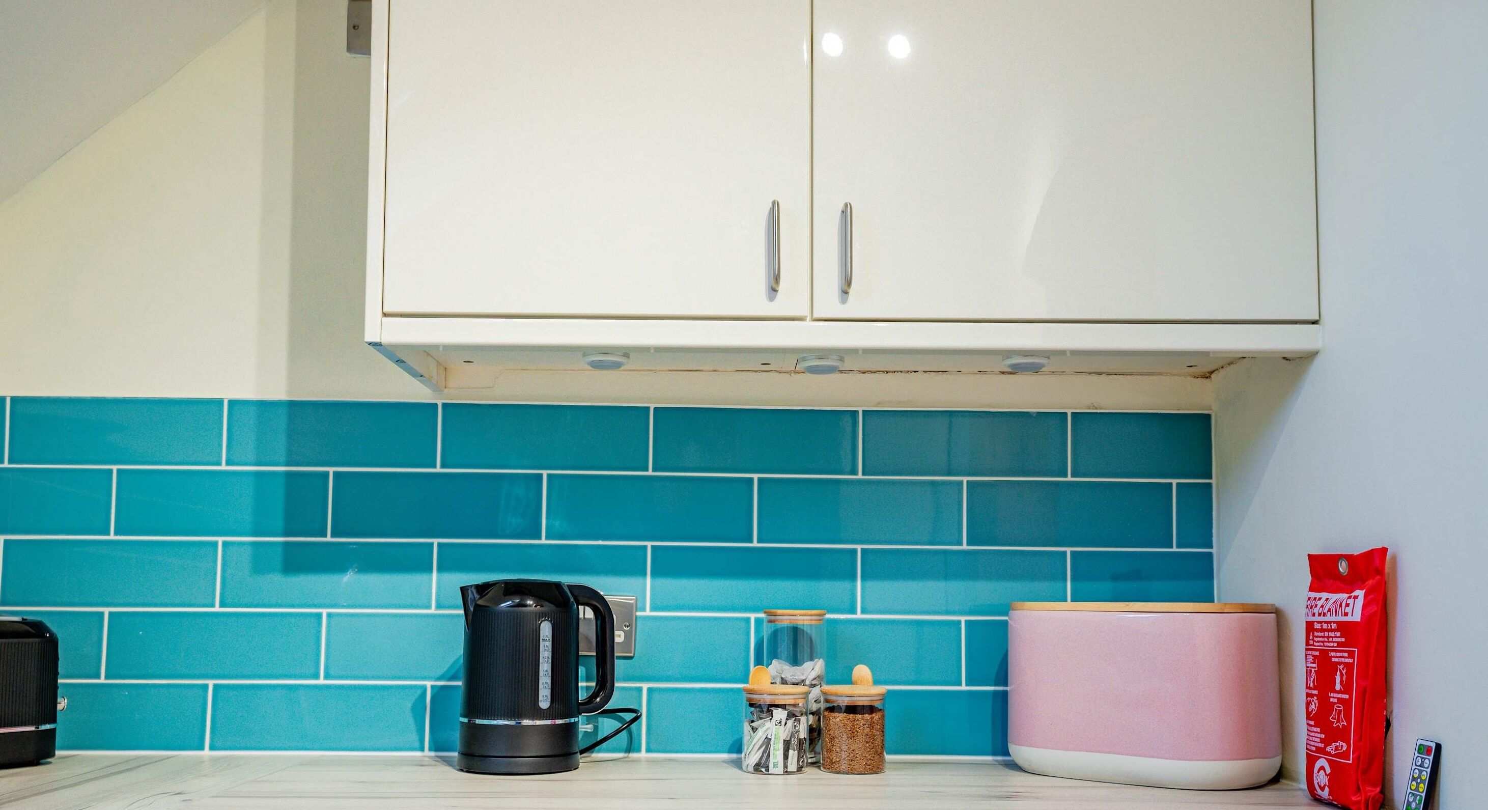 A kitchen with teal tiles, a black electric kettle, a pink container, and a red bag.