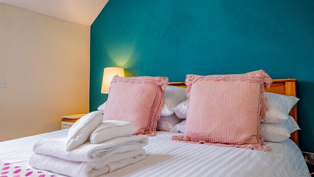 Una cama bien tendida con almohadas rosas y toallas blancas dobladas, una pared de acento color aguamarina y una lámpara de noche.