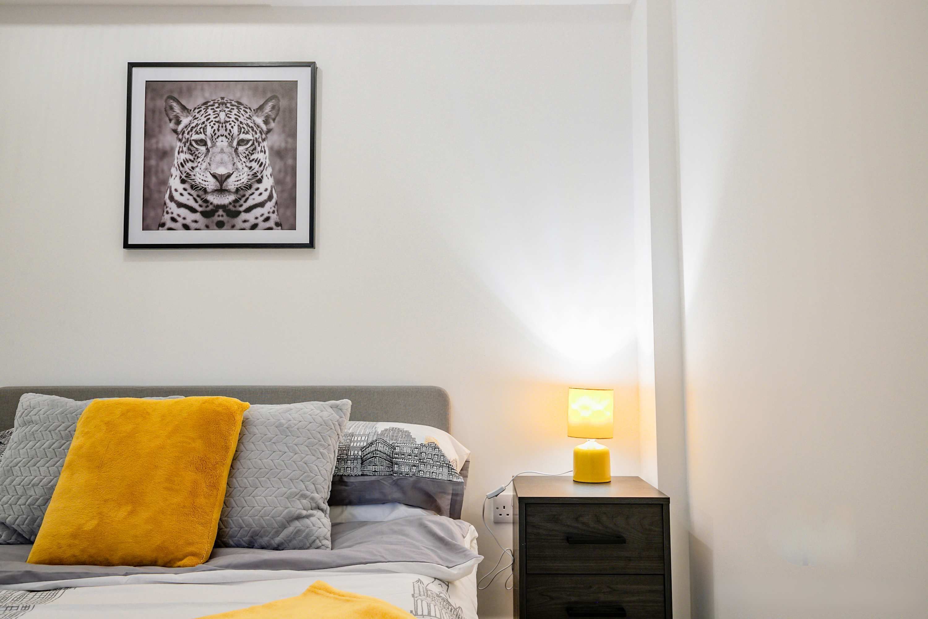 A bedroom with a bed, a bedside table with a lamp, a nightstand, and a framed picture on the wall.