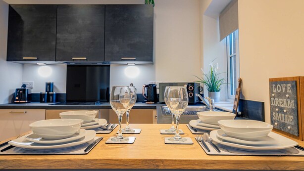 A modern kitchen with dark cabinets, a wooden dining table set with white plates and glasses, and a motivational sign on the wall.