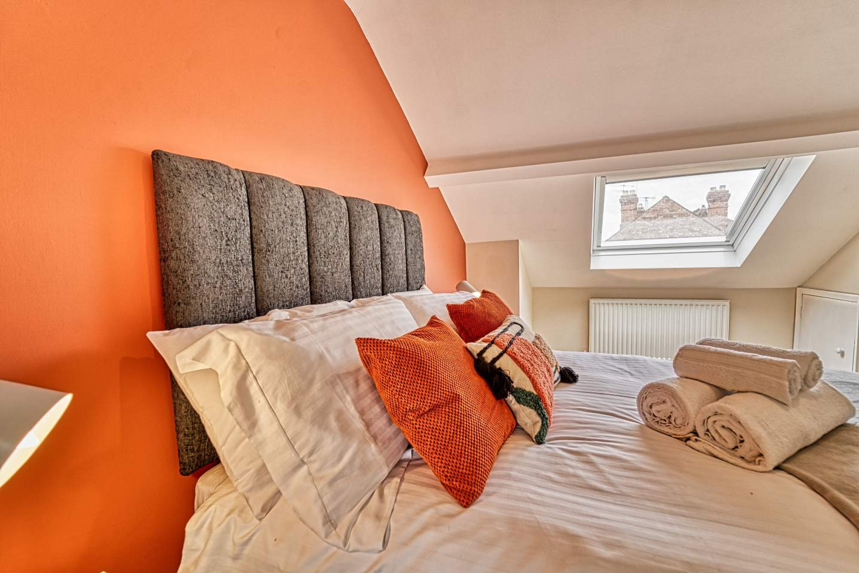 A bedroom with a sloped ceiling, a large window, and a bed with multiple pillows and rolled towels.