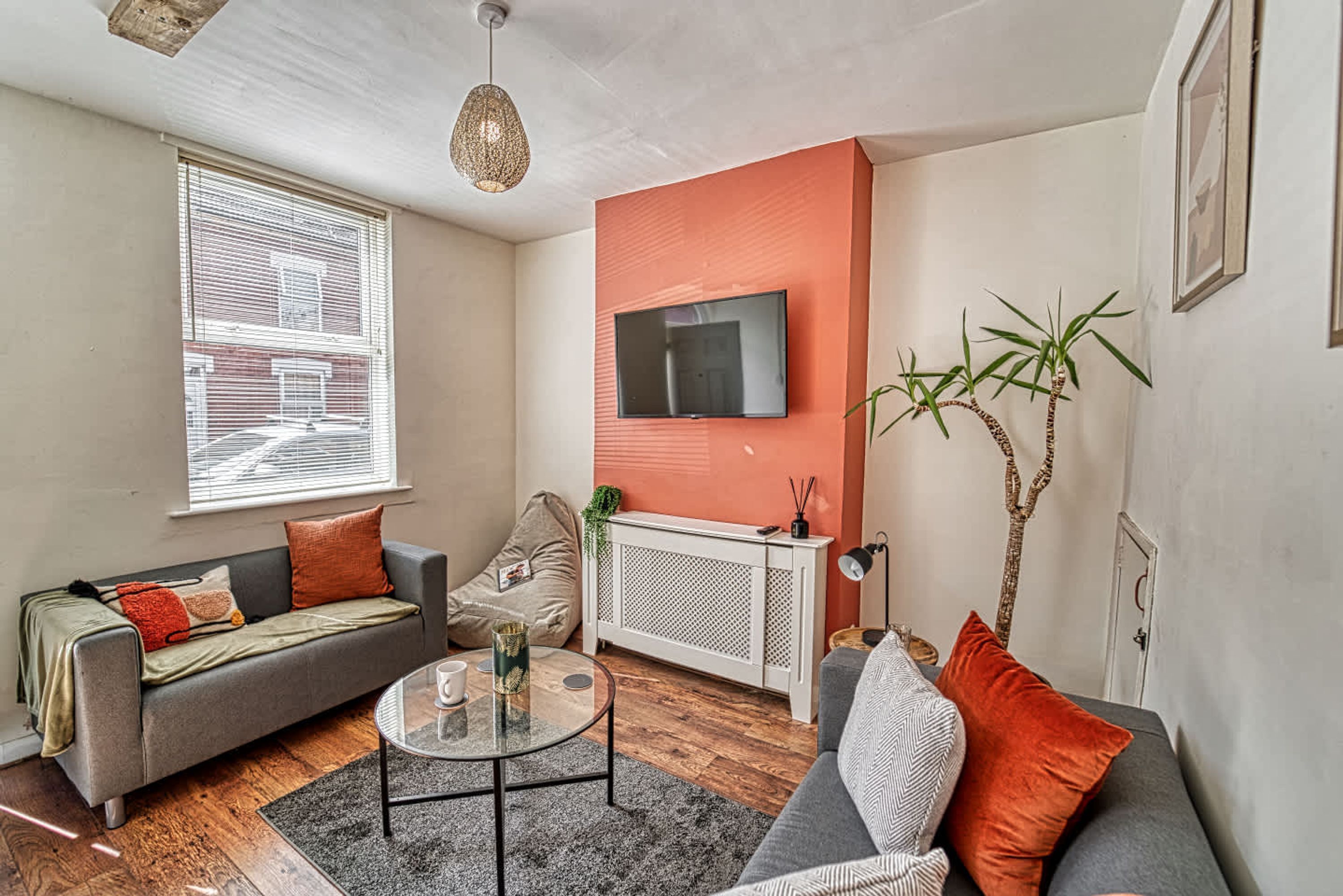 A living room with a sofa, a coffee table, a TV mounted on the wall, and a window with blinds.