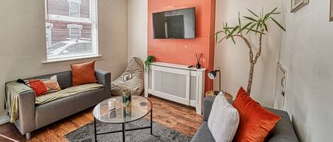 A living room with a sofa, a coffee table, a TV mounted on the wall, and a window with blinds.
