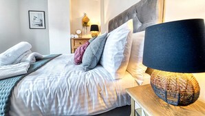 Un lit bien fait, avec une tête de lit grise, une lampe de chevet et un tableau encadré au mur.
