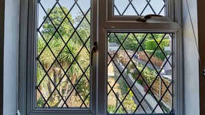 Une fenêtre ornée d’une grille à motifs losangés, donnant sur un jardin où l’on peut admirer diverses plantes et une clôture en bois.