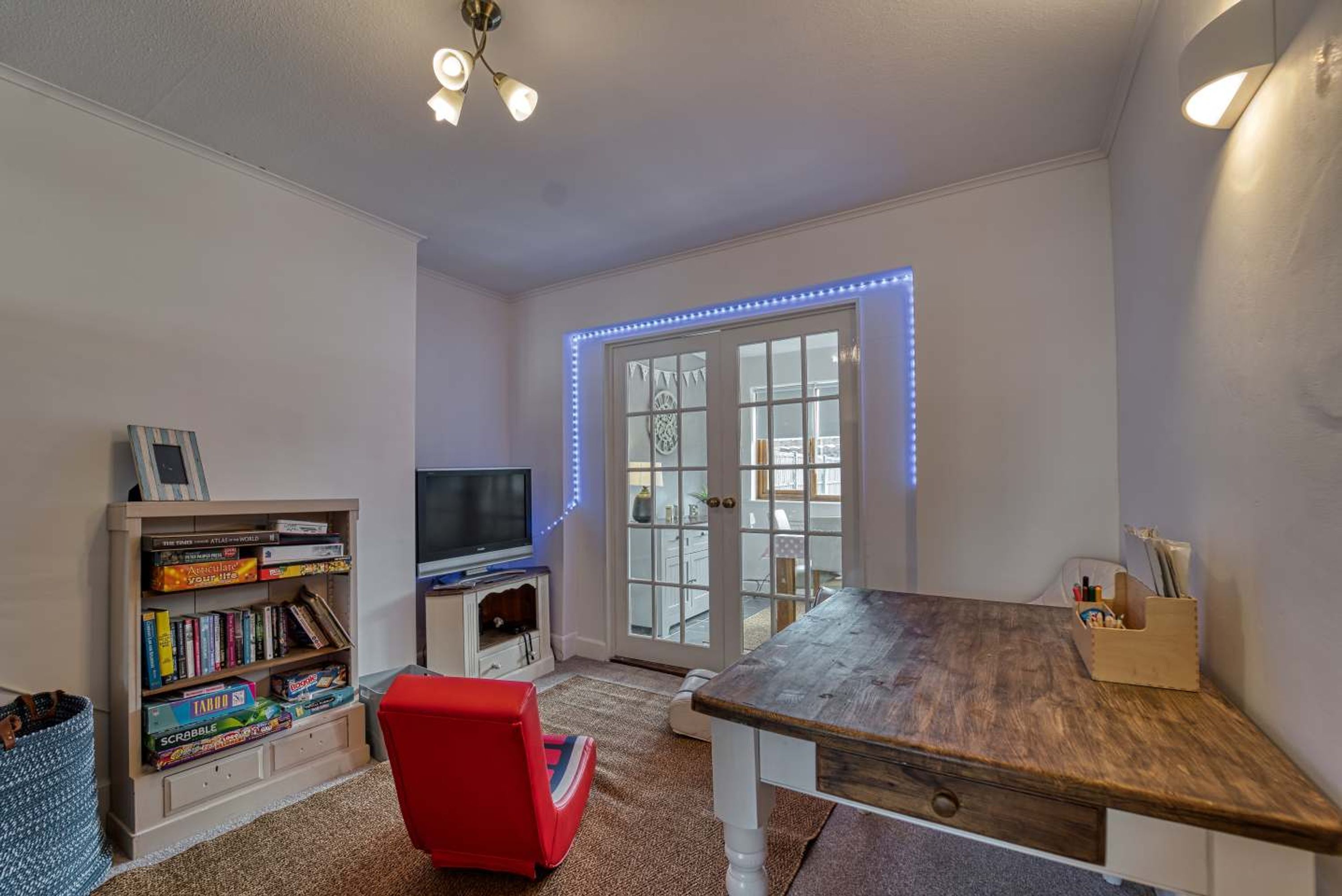 A room with a wooden table, a red chair, a bookshelf with games, a television, and a window with blue LED lights.