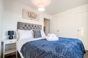 Una cama bien hecha con un edredón azul texturizado, un cabecero gris capitonado y una pared blanca con una obra de arte floral.