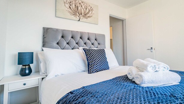 A neatly made bed with a blue blanket, a grey tufted headboard, white and patterned pillows, and a blue lamp on a bedside table. - Host Stay Chestnut Residence (Worcester)
