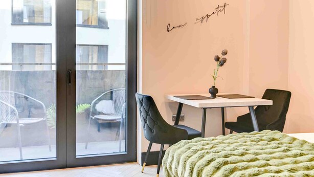 A modern bedroom with a sliding glass door, a bed with a green blanket, two black chairs, a small table with a vase, and a wall with the text 'bon sport.'