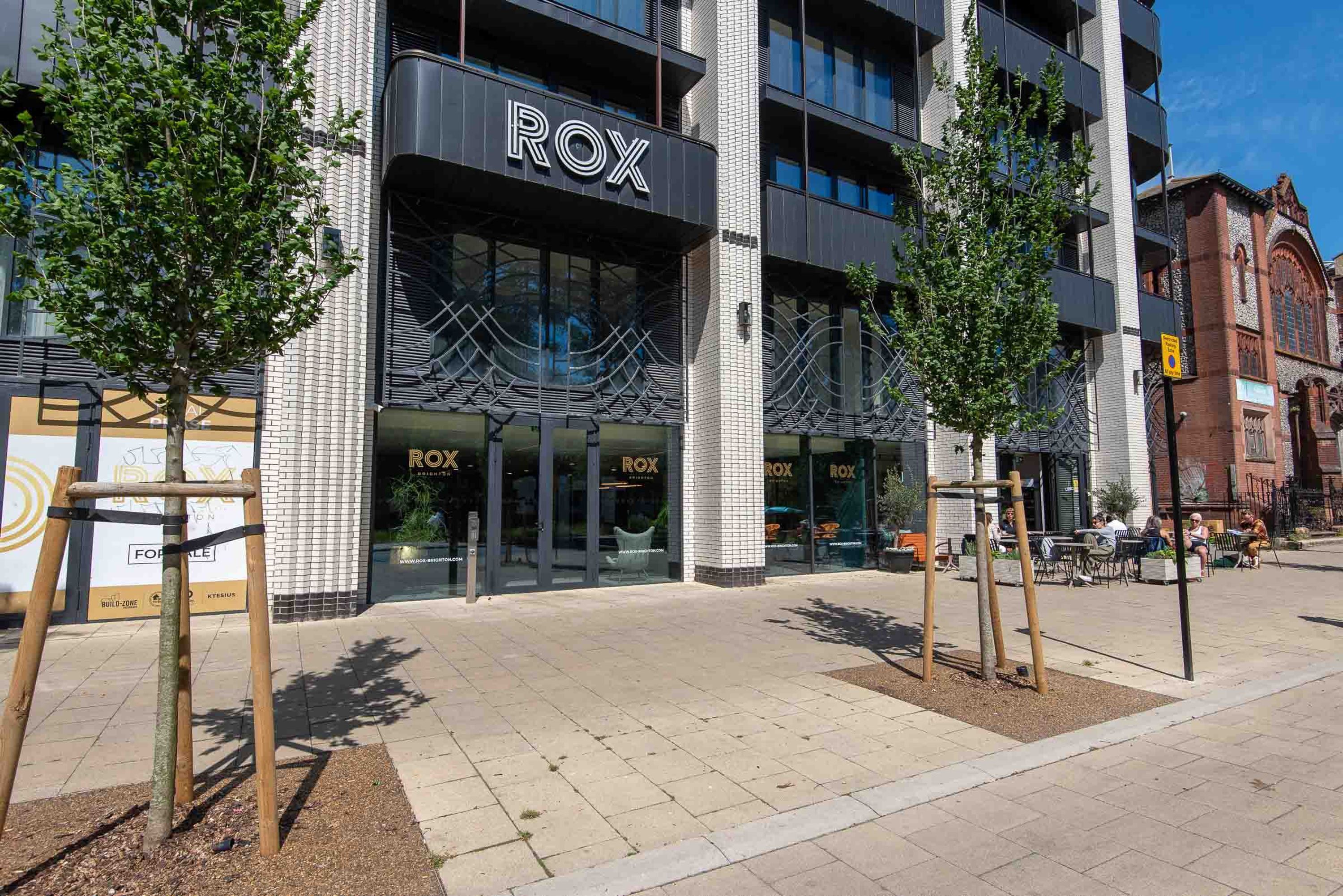 Ένα σύγχρονο κτίριο με την επιγραφή «ROX», ένα πλακόστρωτο πεζοδρόμιο και έναν εξωτερικό χώρο με καθίσματα.