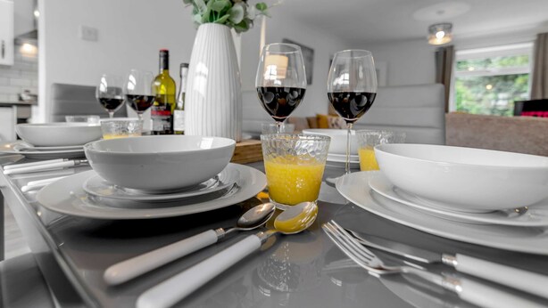 Une cuisine moderne avec des appareils électroménagers blancs, une table à manger dressée avec des assiettes, des bols et des verres contenant du jus d’orange et du vin rouge.