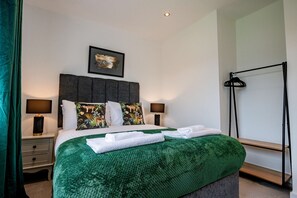 A neatly made bed with a green blanket, two pillows, and a framed picture above it. There are two bedside tables with lamps and a black stand with clothes on it. - Host Stay Carlisle Road House (Worcester)