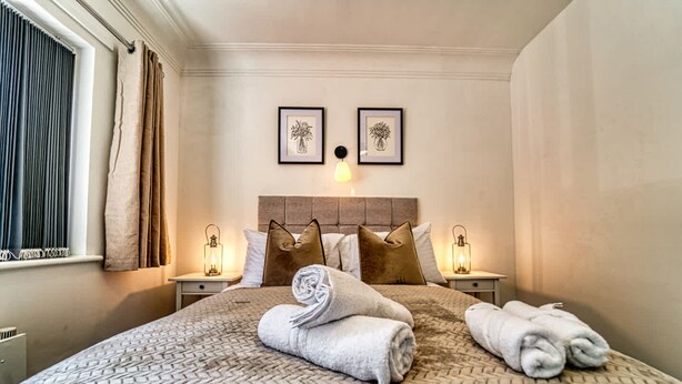 Ein ordentlich gemachtes Bett mit einer gemusterten Tagesdecke, zwei Kissen und gerollten Handtüchern. Zwei Nachttische mit Lampen und zwei gerahmte Bilder an der Wand.