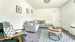Ein modernes Wohnzimmer mit einem grauen Sofa, einem grünen Sessel und einem Couchtisch. An der Wand hängen gerahmte Bilder und im Hintergrund ist eine Tür zu sehen.