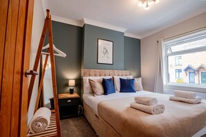 Una cama bien tendida con cabecera, una escalera de madera, un cuadro en la pared y una ventana con vista a edificios.