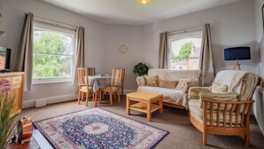 Ein gemütliches Wohnzimmer mit einer Couch, Sesseln, einem Couchtisch und einem Teppich. Es gibt Fenster mit Vorhängen, eine Uhr an der Wand und eine Stehlampe.