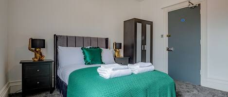 A neatly made bed with a green blanket, two bedside tables with lamps, a wardrobe, and a door with a lock.