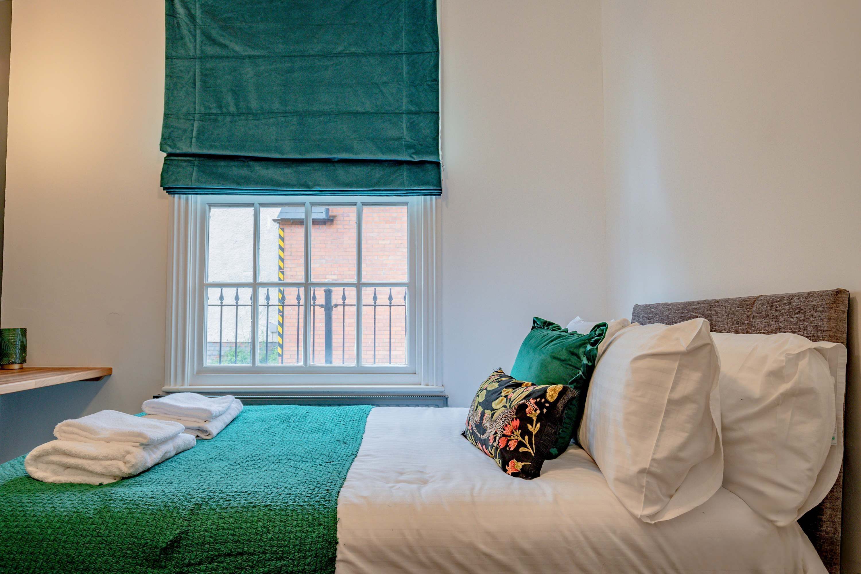 A neatly made bed with white linens, a green throw blanket, and decorative pillows. A window with a view of a brick building is visible.