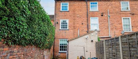 A narrow alleyway with a brick wall on one side and a wooden fence on the other. There are two brick buildings with windows and a door, and a small garden area with a hose and a green bag.