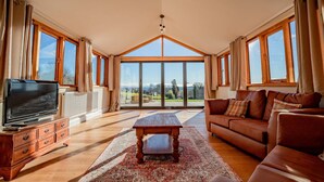 A spacious living room with a large TV, wooden furniture, and a view through large windows. - Host Stay Upperley Barn (Bromyard)