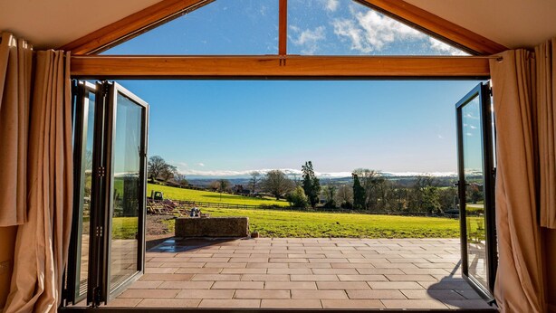 A room with a large glass door leading to a patio with a view of a green landscape and distant mountains. - Host Stay Upperley Barn (Bromyard)