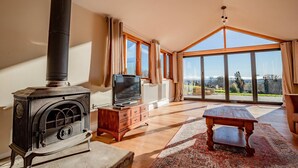 A living room with a wood-burning stove, a television on a wooden cabinet, and a large window offering a view of the outdoors. - Host Stay Upperley Barn (Bromyard)