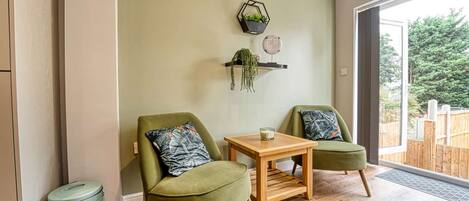 A room with two green armchairs, a small wooden table, and a trash bin. There are two wall-mounted shelves with plants and decorative items.